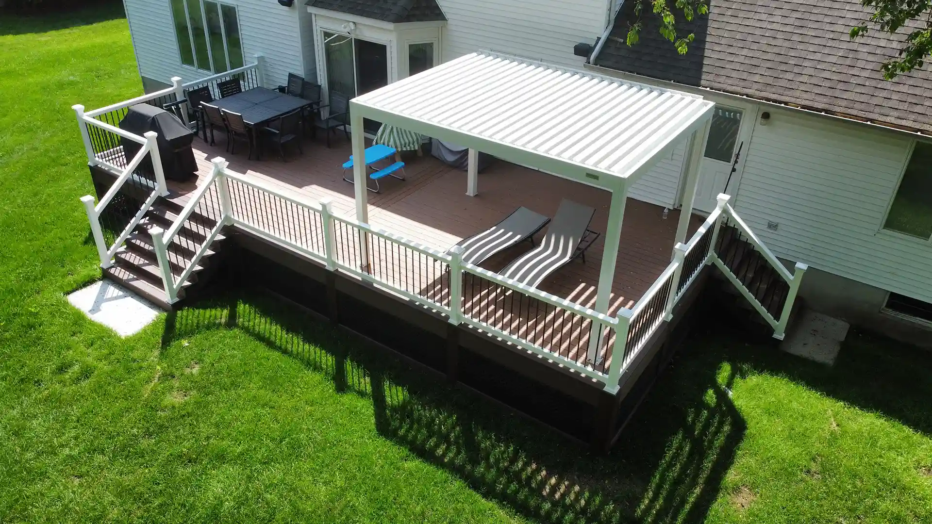 covered porch vs screened porch Photo of a composite deck with white railings and brown flooring. The deck features a white louvered pergola, a dining set, and two lounge chairs.